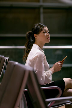 Asian woman sitting on chairs in airport lounge holding smartphone wearing business attire