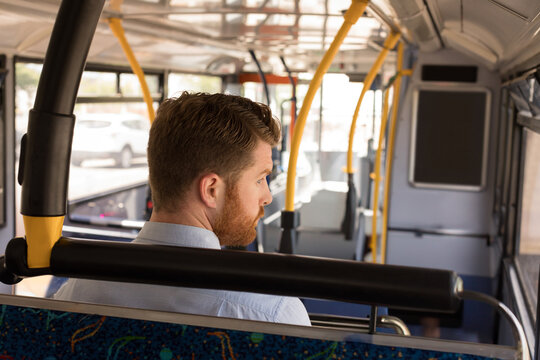 Red-haired passenger sitting on bus seat with yellow poles, sunlit window and parked car outside