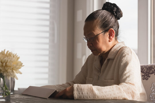 Senior Asian woman reading book wearing glasses at sunlit table with vase of flowers, copy space