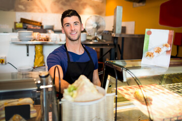 Man in his twenties wearing apron working behind deli counter with glass display case, menu cards