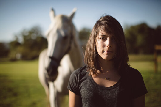 Woman standing in sunlit farm pasture with white horse behind and showing pendant necklace - Powered by Adobe