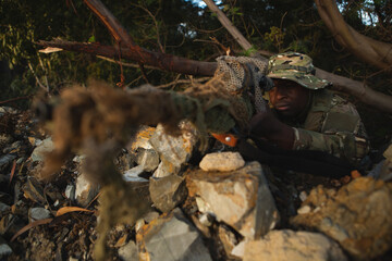 African American man soldier in camo uniform aiming sniper rifle in woods behind rocks, copy space