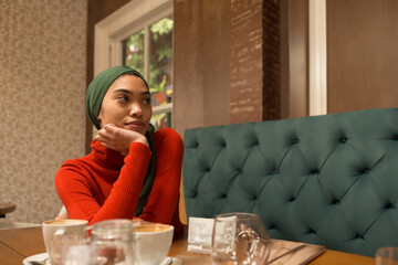 Woman wearing green headscarf and red sweater gazing at cafe table with cappuccino, copy space