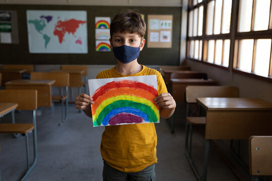 Boy wearing navy face mask holding rainbow painting in classroom by world map poster