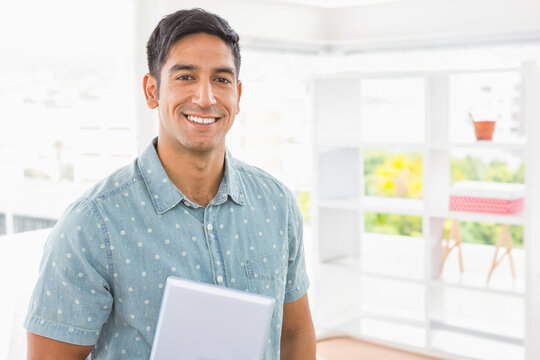 Indian man smiling while holding laptop near office shelves with plant and box, copy space