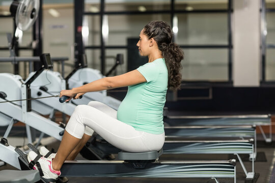 Pregnant woman rowing on machine pulling handle toward torso in fitness center with windows and fan