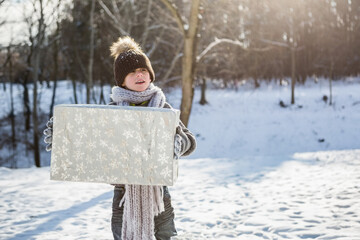 Boy aged seven standing in snowy woodland clearing wearing padded coat and holding silver gift box