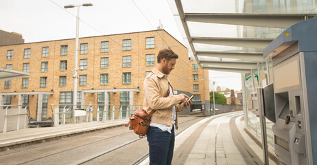 Mid adult male using ticket kiosk on tram platform holding smartphone wearing satchel, copy space
