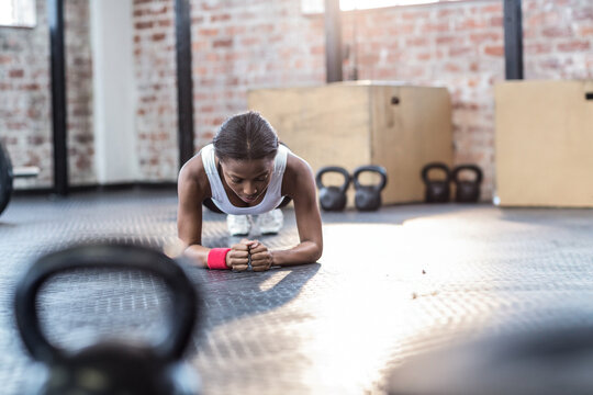 African American woman holding forearm plank on mat in gym with kettlebells and plyo boxes