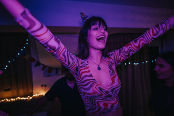 A group of friends enthusiastically celebrate a birthday at home, surrounded by colorful lights and...