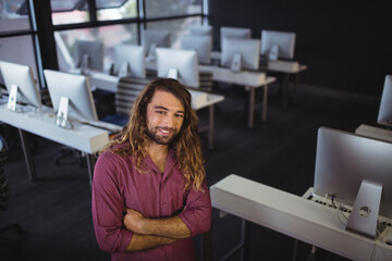 Male student standing arms crossed in computer lab with monitors, keyboards wearing maroon shirt