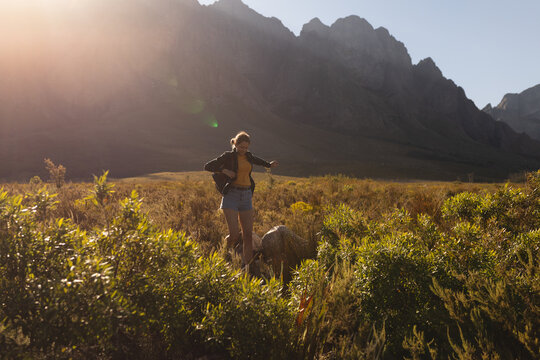 Female hiker standing on rock outcrop in grassland at mountain base watching light-coated dog
