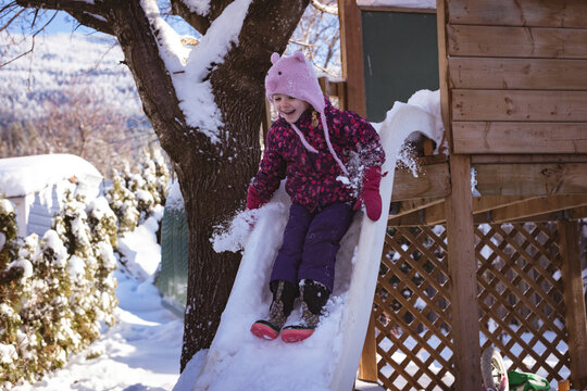 Girl is sliding down snow-covered plastic slide on playhouse in backyard with shrubs and mountains