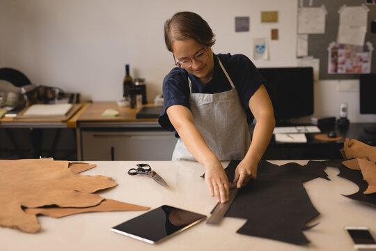 Woman wearing gray apron measuring cutting black brown leather hides with ruler at workshop bench - Powered by Adobe