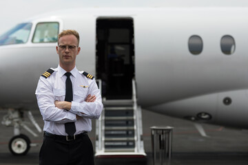 Male pilot standing with arms crossed on tarmac by private jet stairs, copy space