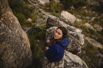 Female hiker perching on boulder in blue jacket with dark backpack, looking upward on slope