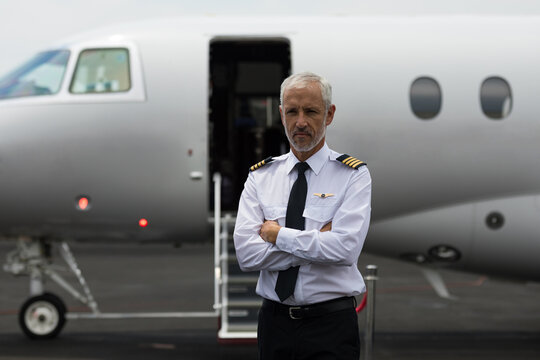 Senior male pilot standing with arms crossed on ramp before private jet wearing epaulets and tie