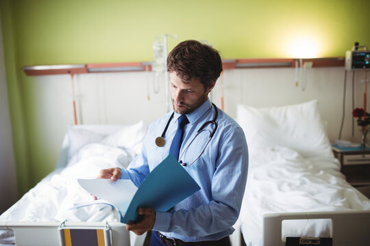 Mid adult male doctor wearing stethoscope reviewing patient file beside hospital bed, IV stand
