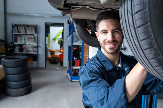 Male mechanic working under vehicle on hydraulic lift in garage with stacked tires, copy space