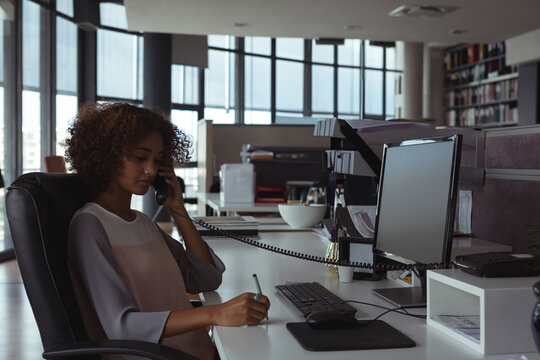 African American woman holding phone and writing notes at office desk with monitor, copy space
