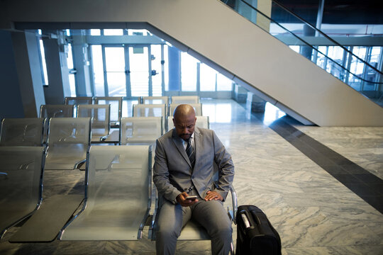 African American man in suit sitting at escalator in airport holding smartphone before glass doors