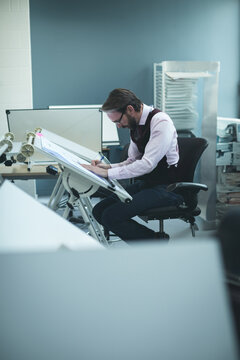 Designer sketching blueprint on tilted drafting table next to computer monitors in design studio