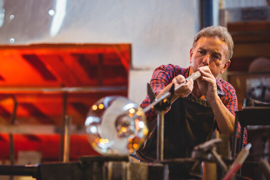 Senior man in dark apron, holding blowpipe shaping molten glass at workshop workbench, copy space
