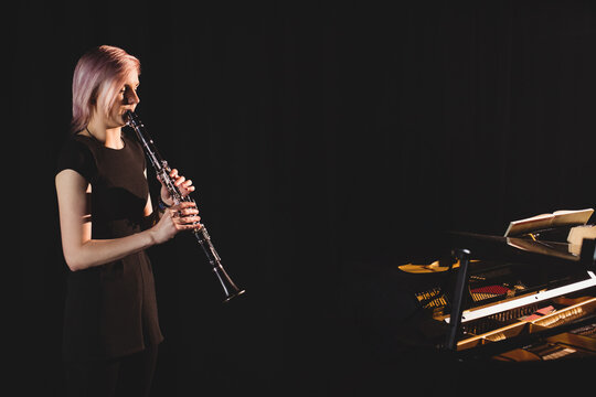 Female musician playing clarinet under spotlight on lit stage beside grand piano with sheet music