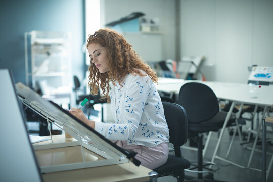 Female architecture student sketching on angled drafting table in design studio with green marker