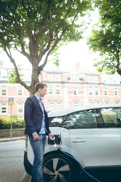 Man connecting blue cable to white electric hatchback by stone wall on leafy urban street