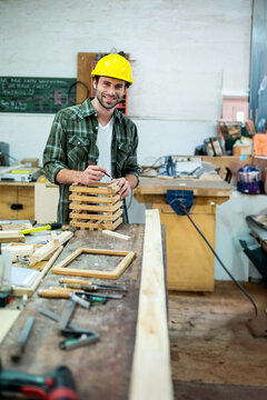 Male carpenter wearing yellow hard hat aligning wooden slat assembly at workbench with tool racks