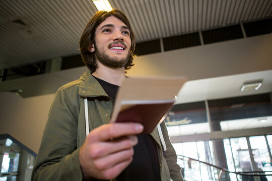 Male traveler wearing green jacket standing at airport concourse holding passport and boarding pass
