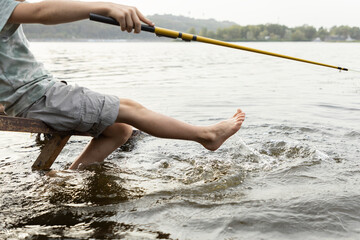 bare feet of a child with a fishing rod splashing over the water while enthusiastically fishing on...