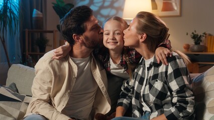 Caucasian happy family at home parents on sofa in evening talking surprised smiling about suddenly daughter child play jumping from behind couch playful kid mother and father kissing girl on cheeks