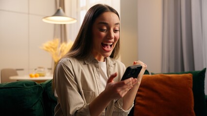 Homeowner young girl woman lady female sitting on couch at home living room mobile phone win game online betting achieve victory e-commerce discount in net virtual triumph celebrate winning success