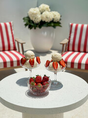 Summer terrace or balcony.  Two glass dessert bowls filled with creamy vanilla ice cream wit fresh strawberries on white  table and two  comfortable armchairs with red and white striped  cushions.