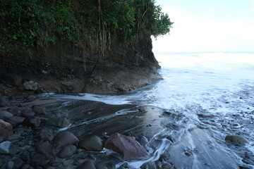 Ocean waves flow over dark rocks and black sand at the base of a lush, cliffside tropical coast...