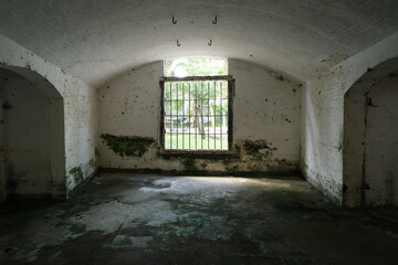 A spacious, arched historical prison cell with barred windows and peeling white walls, showing age,...