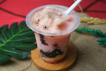 Traditional Es Doger dessert in a clear plastic cup with shaved ice, black glutinous rice, avocado, fermented cassava, and coconut milk, served with a spoon.