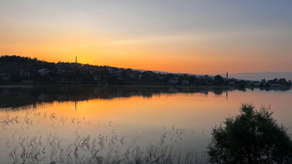 Fototapeta premium It shows a village surrounded by soft orange sunset lights on a calm lake. The reflection of the sky and the village silhouette on the lake creates a peaceful and tranquil atmosphere.