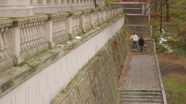 A young couple descends a stone staircase in a city park during autumn. They walk together down the path, with a large historic building and a stone balustrade visible in the scene.