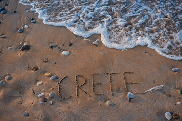 Word Crete written on sand with a feather and sea foam nearby. The concept of tourism on this island