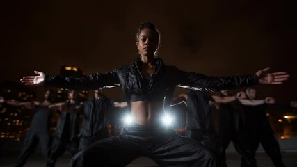 Urban dance crew performs choreography on a rooftop at night in front of city skyline, spotlight on the lead female dancer