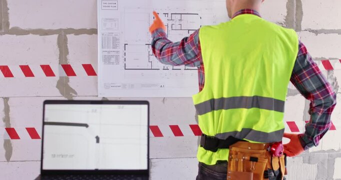 Specialist studies building layout before starting renovation. Engineer in workwear looks at floor plan standing behind laptop with professional program on screen