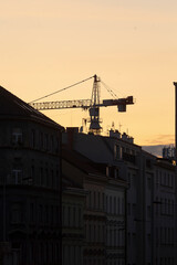 Fototapeta premium Silhouette of a construction crane against a sunset sky, behind urban buildings. Represents city development and construction activities