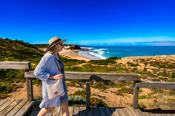 Mature woman in summer hat tourist walking on wooden stairs overlooking at Monte Clerigo beach on Algarve coast on sunny spring day. Side view