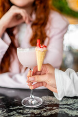 A close-up of a hand holding a pink cocktail glass with a small ice cream cone attached to the rim, set against a blurred background with warm tones. stylish presentation adds a unique touch to drink