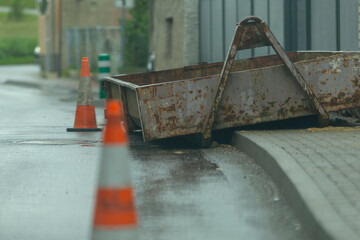 An abandoned container filled with construction debris is sitting on a wet, slick road, surrounded...