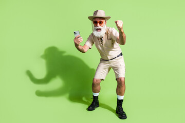 Photo of excited adventurer senior man use cell gadget raise fists up winning casino treasure isolated green color background