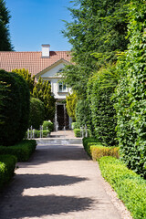 Symmetrical garden alley with trimmed hedges, decorative fountain, and a cozy house in the background on a sunny summer day.